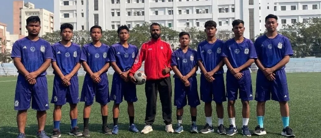 Eight ICFAI FC players in royal blue kit lined up on the pitch, with the club In-Charge holding a football in the centre